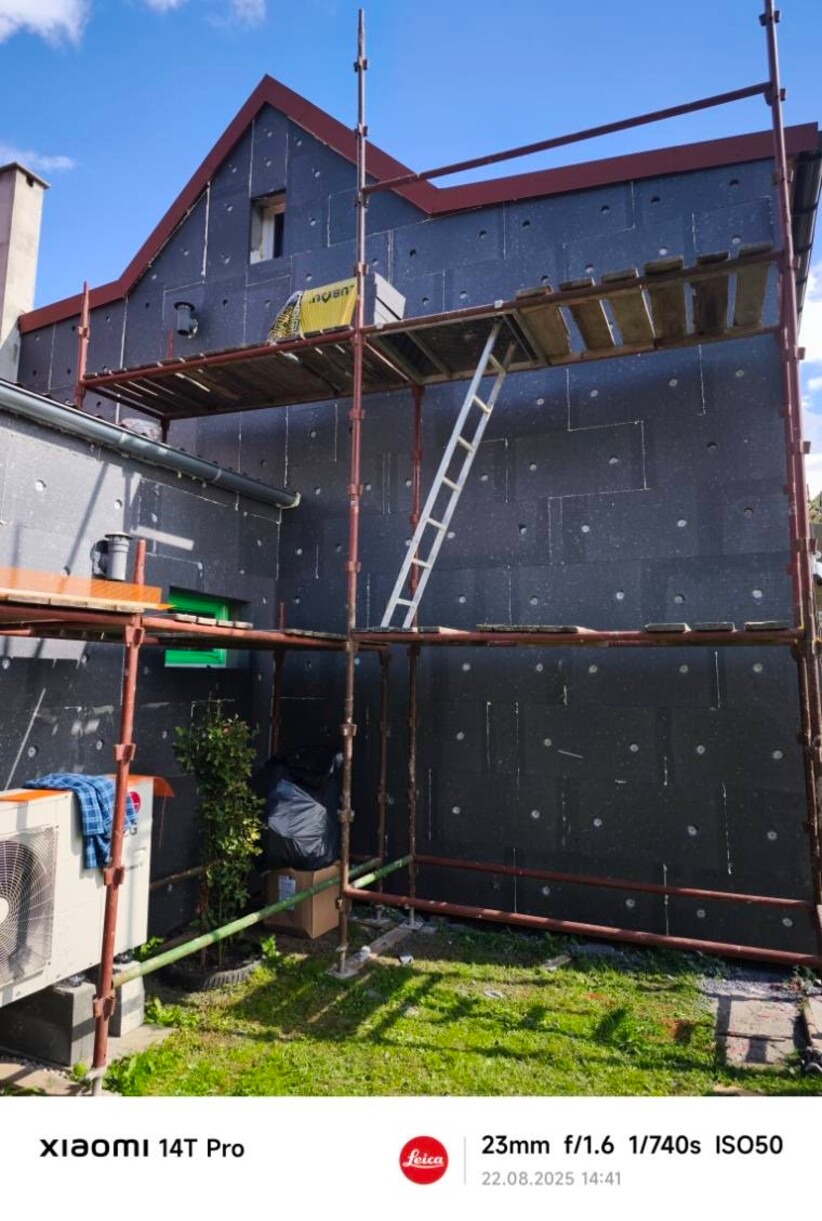 Dom w trakcie ocieplania, widoczne rusztowania i czarne płyty izolacyjne na elewacji. Na pierwszym planie trawnik i klimatyzator.