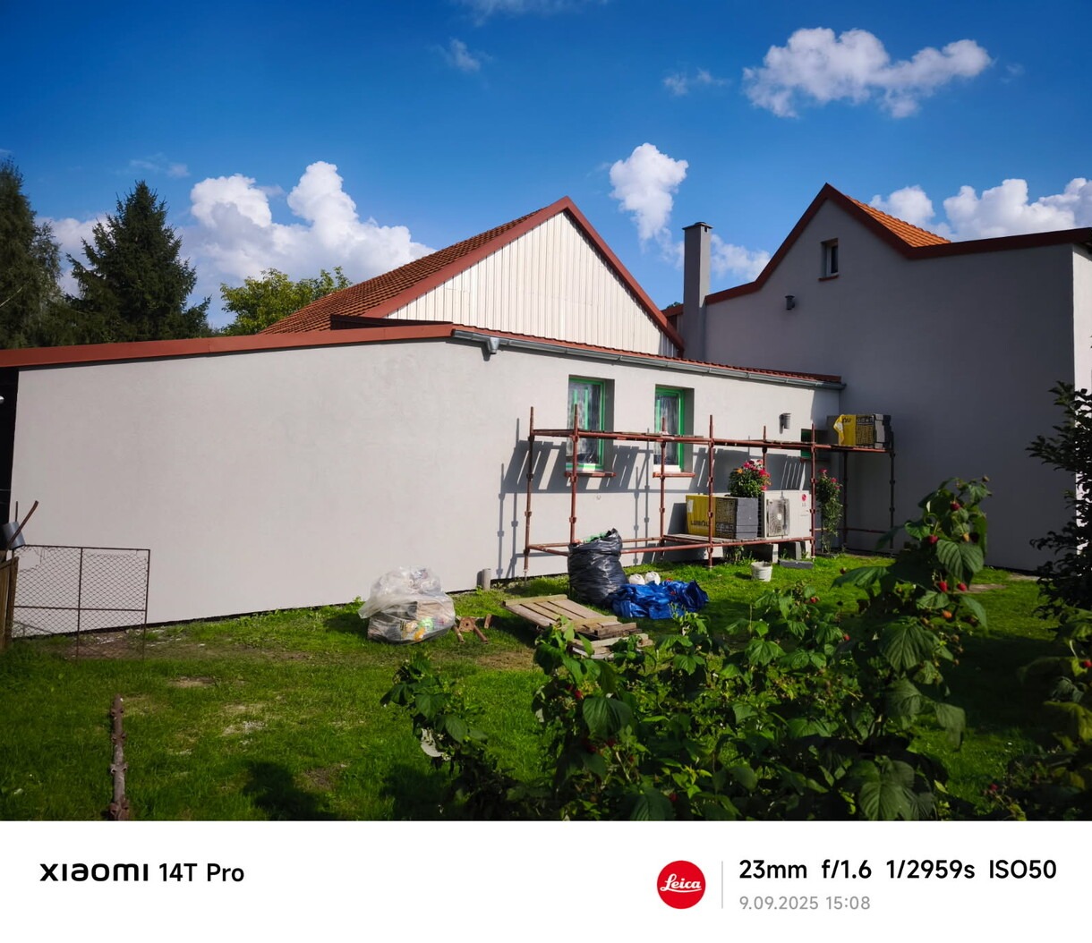 Jasna elewacja domu jednorodzinnego po malowaniu, z widocznymi rusztowaniami i elementami przygotowania do prac. Błękitne niebo z chmurami w tle.