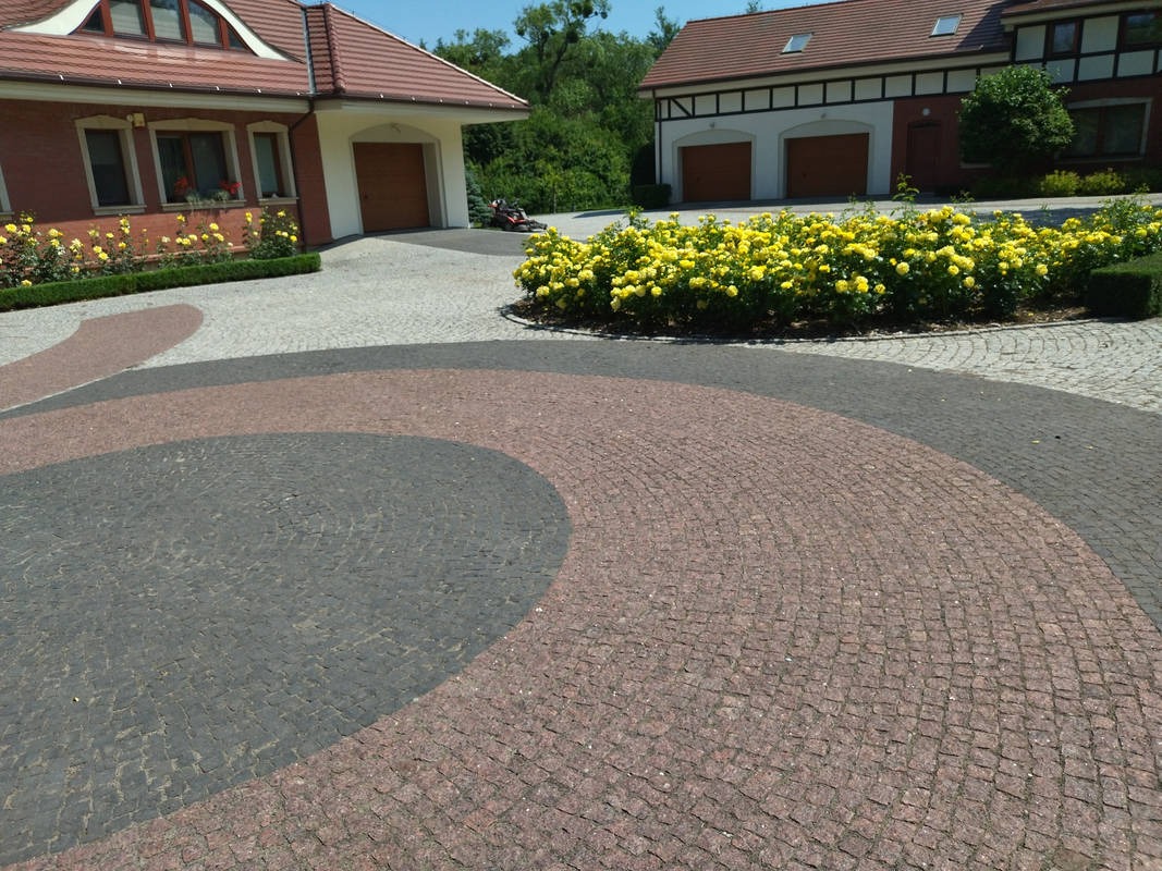 Kolorowa kostka brukowa tworzy wzór na podjeździe. Żółte róże zdobią rabaty, a w tle widoczne budynki z garażami i zieleń drzew.