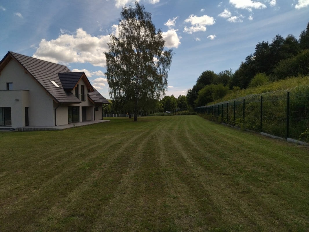 Rozległy trawnik przed nowoczesnym domem z ciemną dachówką, obok wysokie drzewo i ogrodzenie z siatki, błękitne niebo z chmurami w tle. Teren po koszeniu.