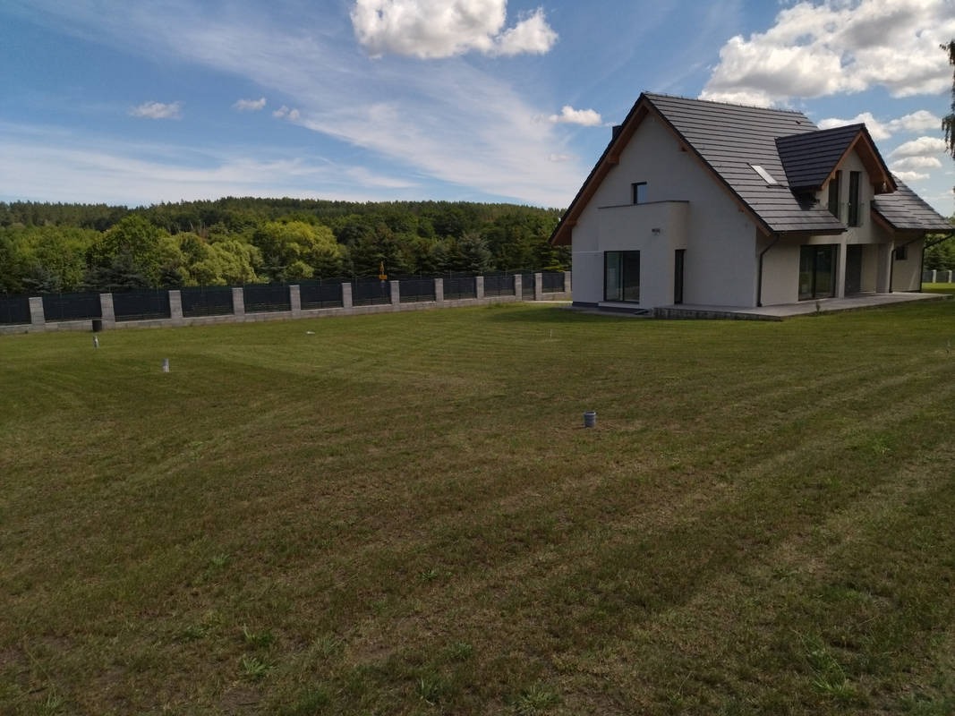 Rozległy trawnik przed nowoczesnym domem z szarą dachówką i ogrodzeniem. W tle widoczny las pod błękitnym niebem z chmurami.