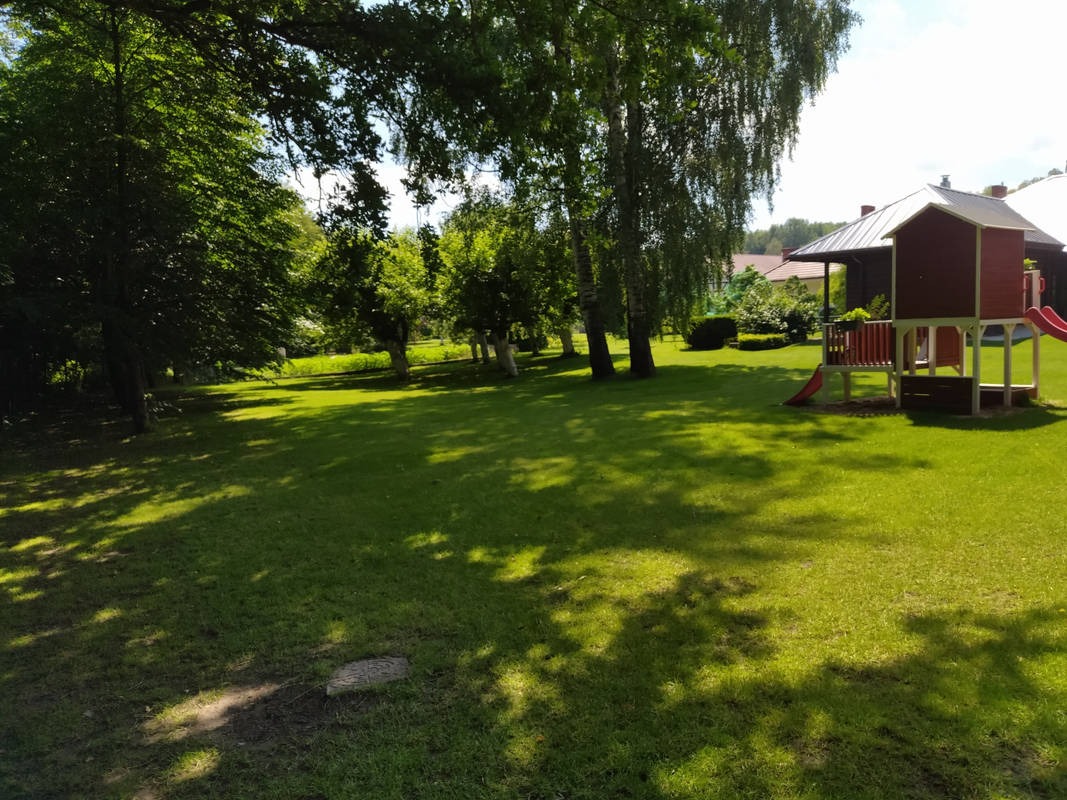 Zadbany trawnik z drzewami i domkiem dla dzieci w słoneczny dzień. Widoczne cienie drzew na trawie. Trawa równo skoszona. W tle zabudowa.