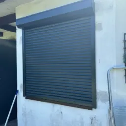 Zamknięta, grafitowa roleta zewnętrzna, zamontowana na elewacji budynku z widoczną fakturą. W tle fragment drzwi i drabina.