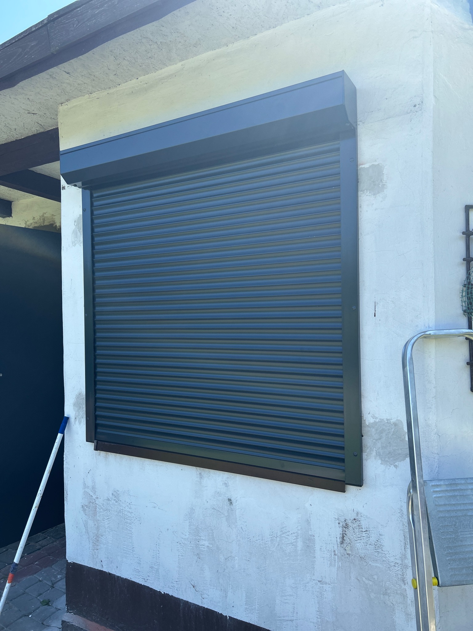 Zamknięta, grafitowa roleta zewnętrzna, zamontowana na elewacji budynku z widoczną fakturą. W tle fragment drzwi i drabina.