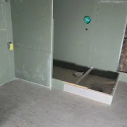 Montaż płyt gipsowo-kartonowych w łazience, widoczna konstrukcja brodzika i instalacja odpływowa. Ściany przygotowane do dalszych prac wykończeniowych.