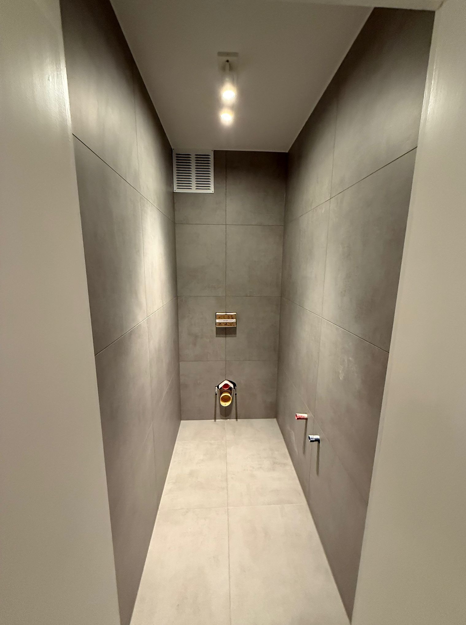 Wnętrze łazienki w trakcie wykańczania, z szarymi płytkami i widocznymi instalacjami wodno-kanalizacyjnymi. Minimalistyczny styl, surowy beton, oświetlenie sufitowe.