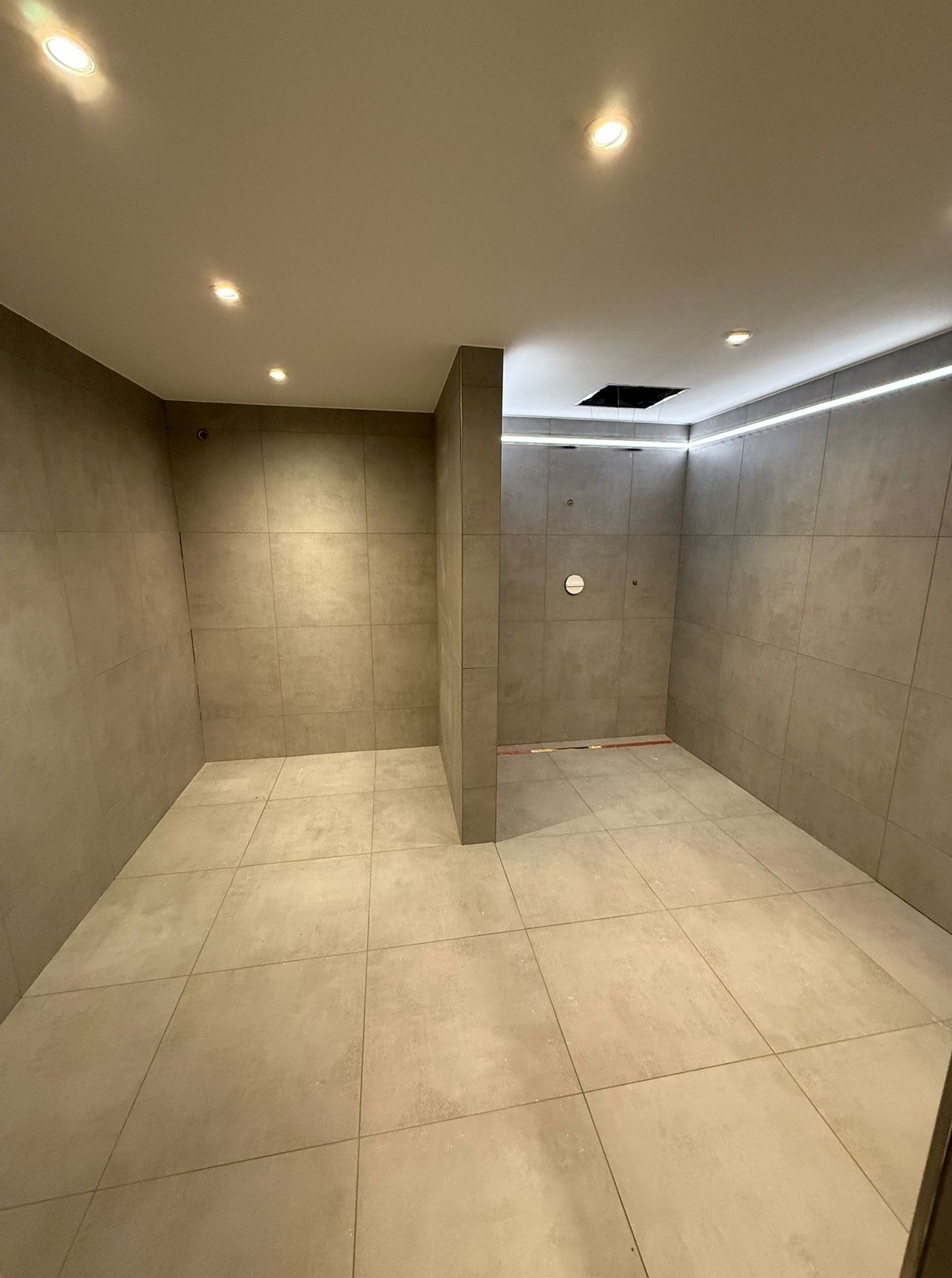 Wykończona, nowoczesna łazienka z beżowymi płytkami, oświetleniem LED na suficie i punktowymi lampami. Widoczny otwór w suficie, prawdopodobnie na wentylator.