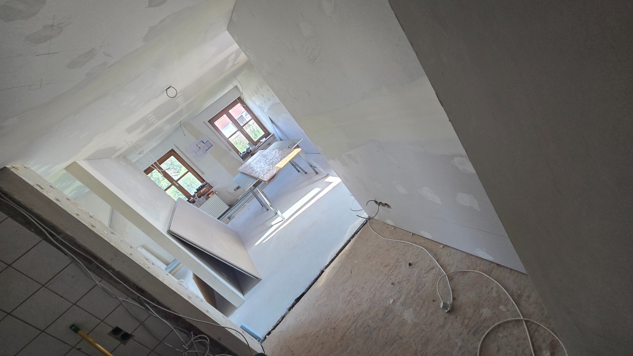 Wnętrze w trakcie szpachlowania ścian i sufitu. Widoczne okna, narzędzia i kable. Prace wykończeniowe w jasnym pomieszczeniu z widokiem na zewnątrz.