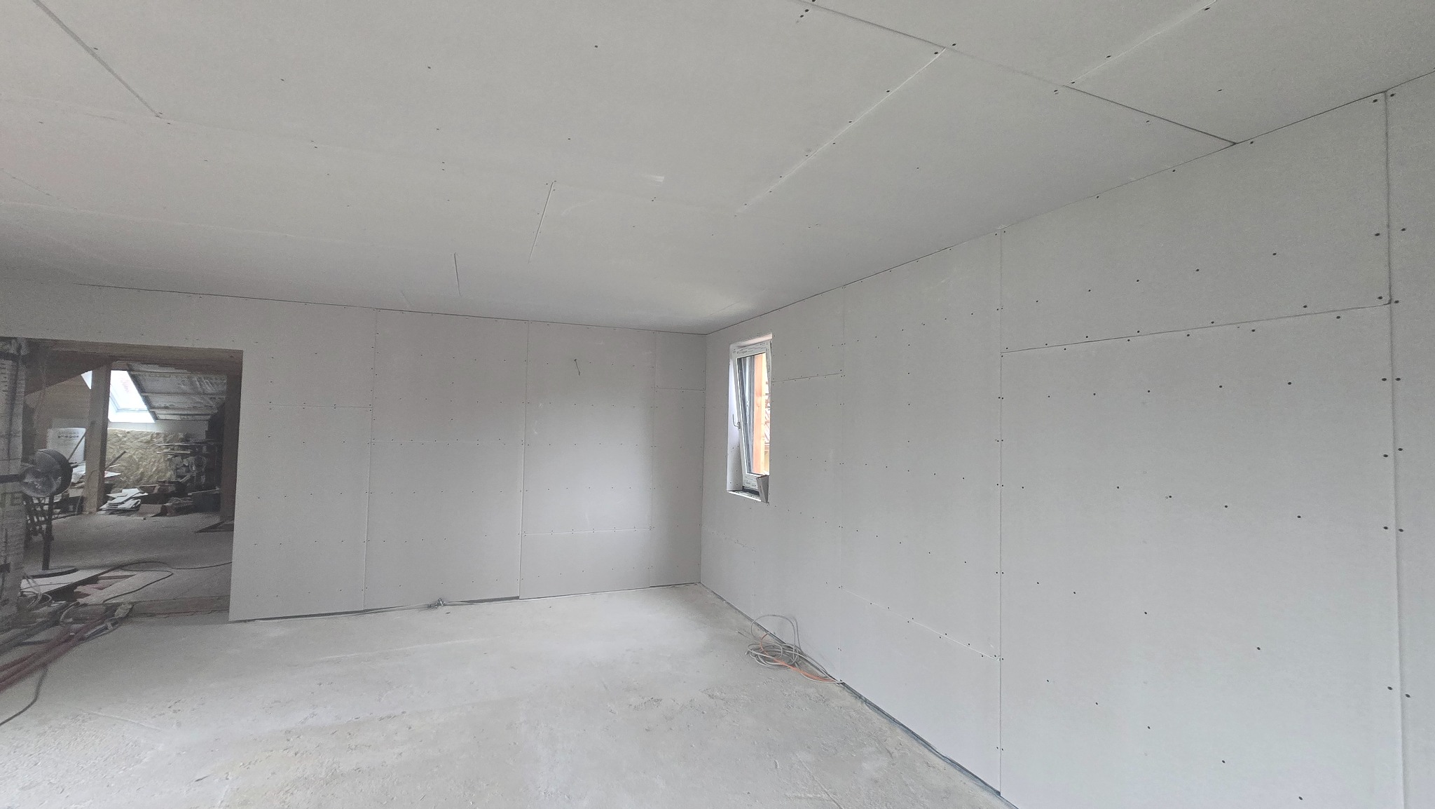 Pokój po montażu płyt gipsowo-kartonowych na ścianach i suficie, widoczne łączenia płyt, okno i przewody na podłodze. Otwór drzwiowy w tle.