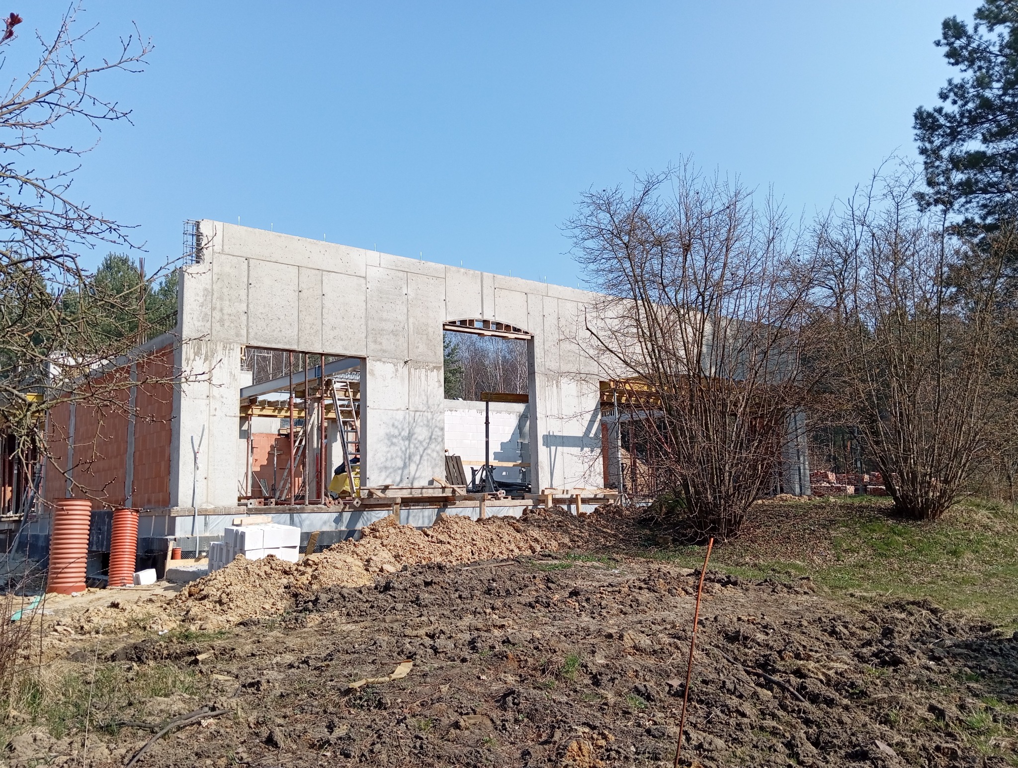 Budowa domu jednorodzinnego w stanie surowym: ściany z betonu komórkowego, widoczne zbrojenia, szalunki, teren budowy z ziemią i drzewami w tle.