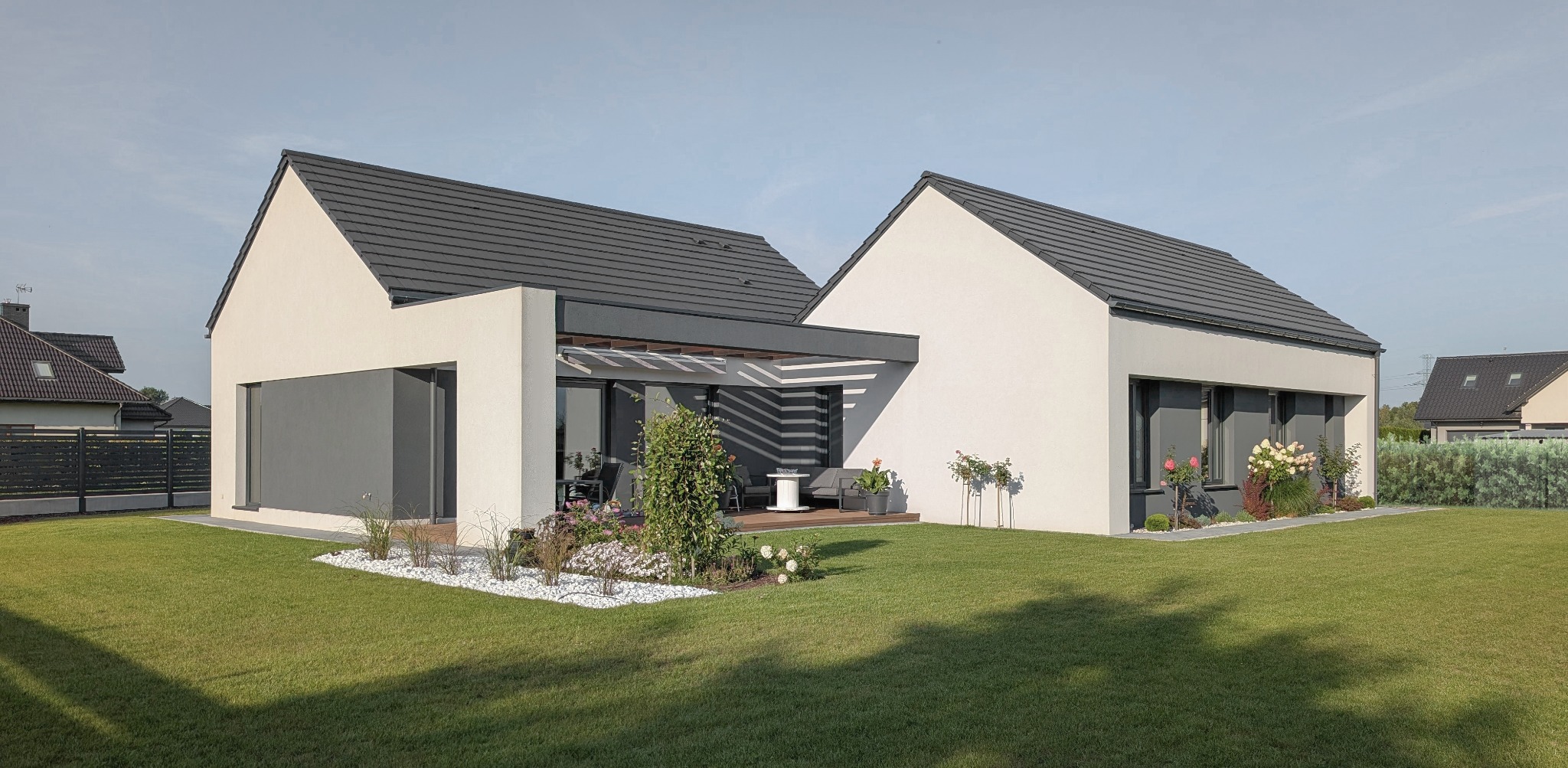 Nowoczesny dom jednorodzinny z płaskim dachem, jasną elewacją i szarymi akcentami. Widoczny taras z pergolą i zadbany trawnik z elementami dekoracyjnymi.