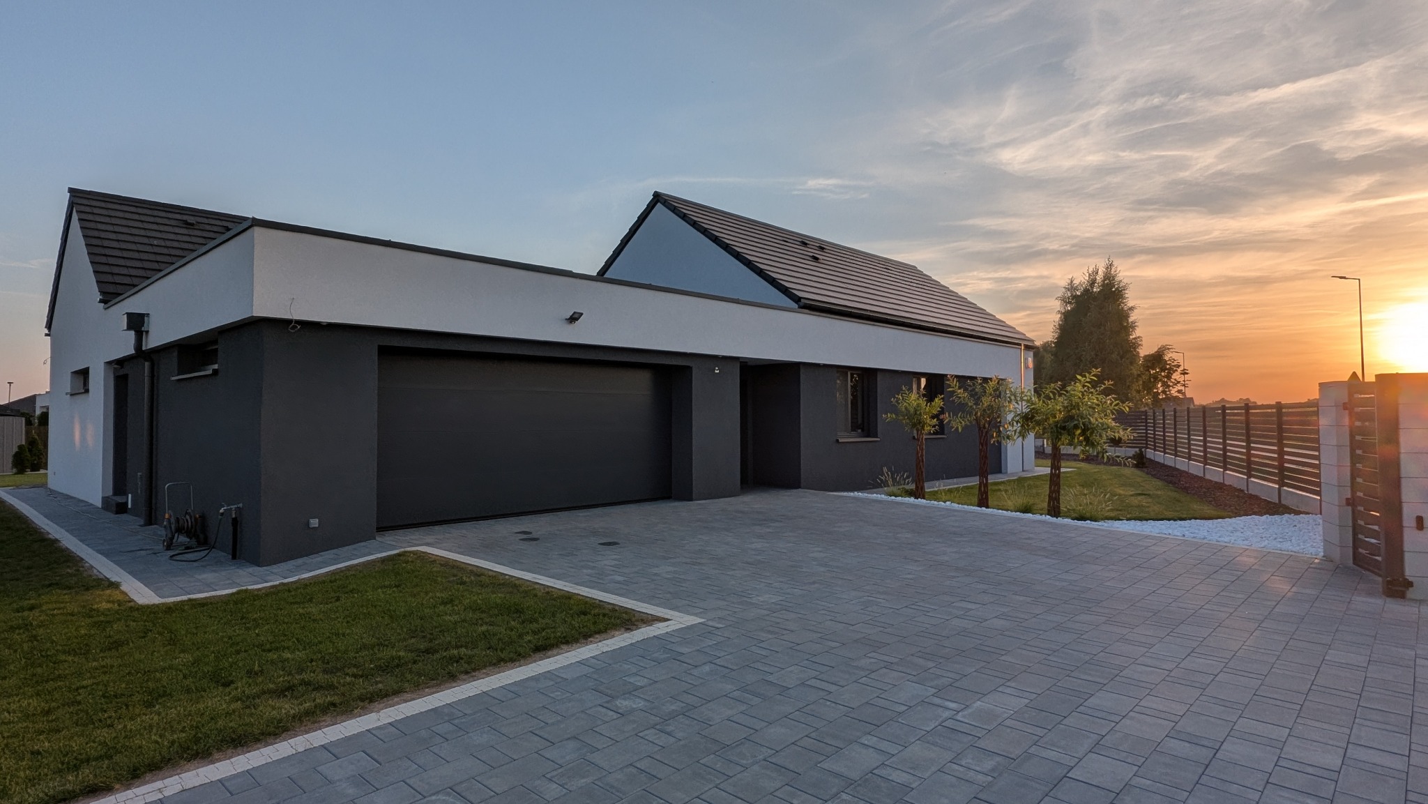 Nowoczesny dom jednorodzinny z garażem i podjazdem z kostki brukowej w odcieniach szarości, oświetlony ciepłym światłem zachodzącego słońca.