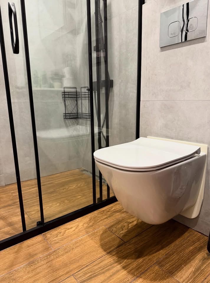 Nowoczesna łazienka z podwieszaną toaletą, kabiną prysznicową z czarnymi ramami i drewnopodobną podłogą. Minimalistyczny design, płytki w odcieniach szarości.