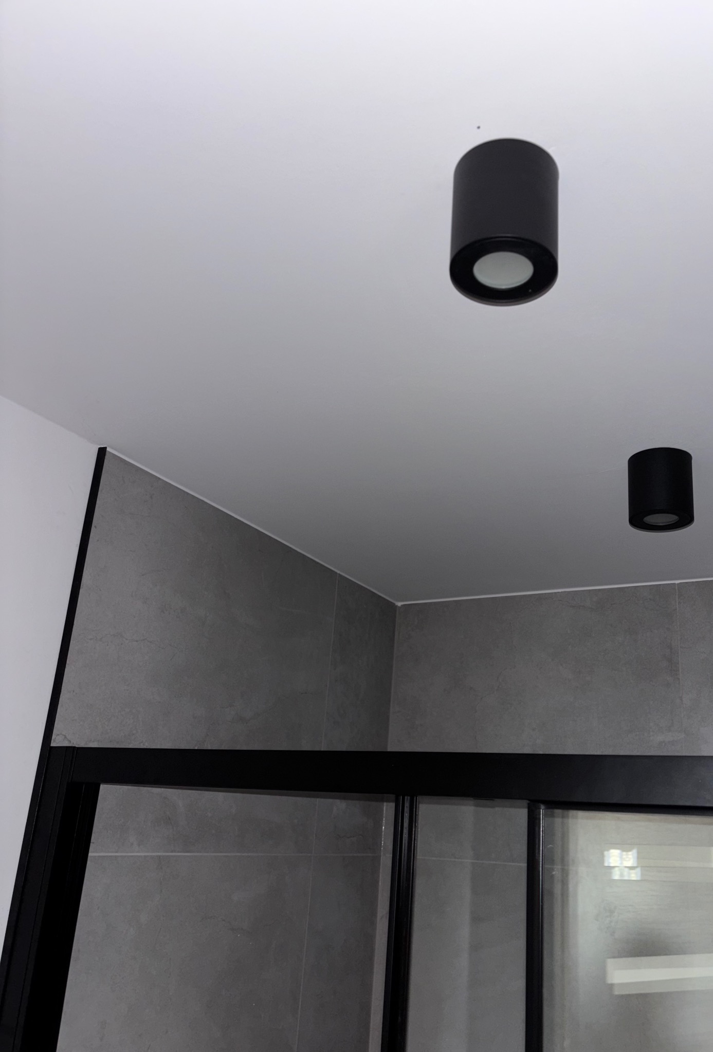 Nowoczesna łazienka z szarymi płytkami, czarną kabiną prysznicową i dwoma okrągłymi, czarnymi lampami sufitowymi. Minimalistyczny design.