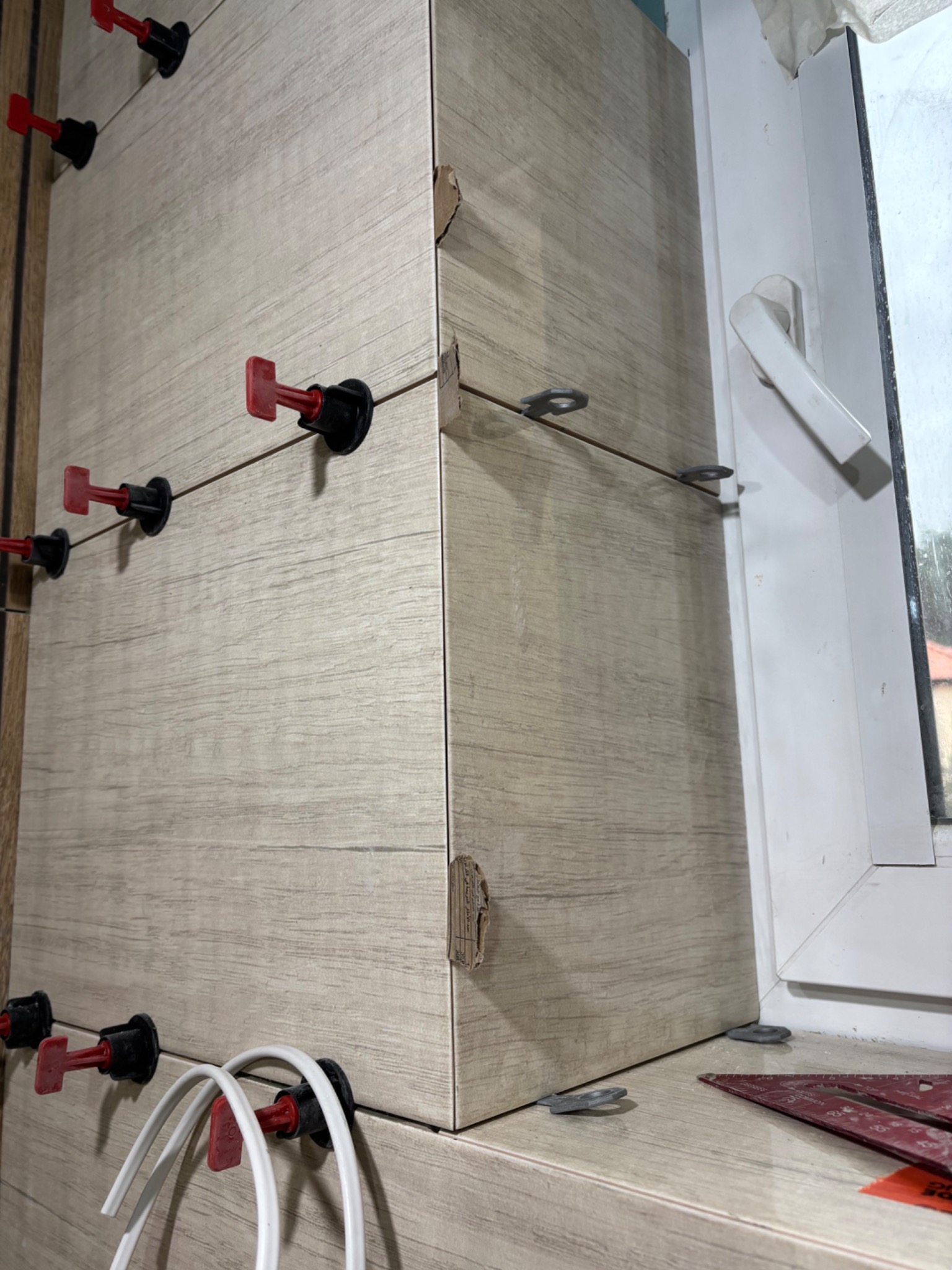 Układanie płytek imitujących drewno przy oknie, z użyciem systemu poziomowania płytek. Widoczne narzędzia glazurnicze i fragment okna.