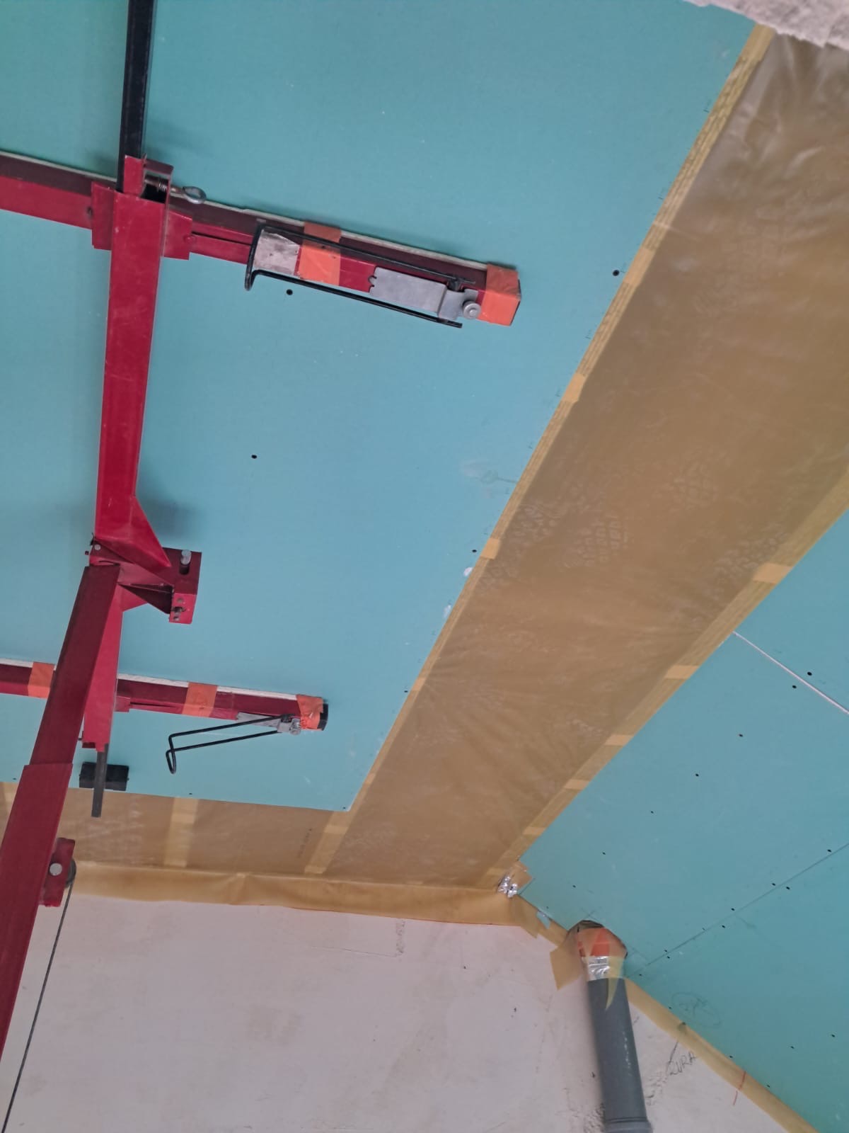 Montaż sufitu podwieszanego z płyt gipsowo-kartonowych. Widoczny stelaż i płyty w kolorze turkusowym, zabezpieczone taśmą. Ujęcie z dołu, widoczny fragment rury.