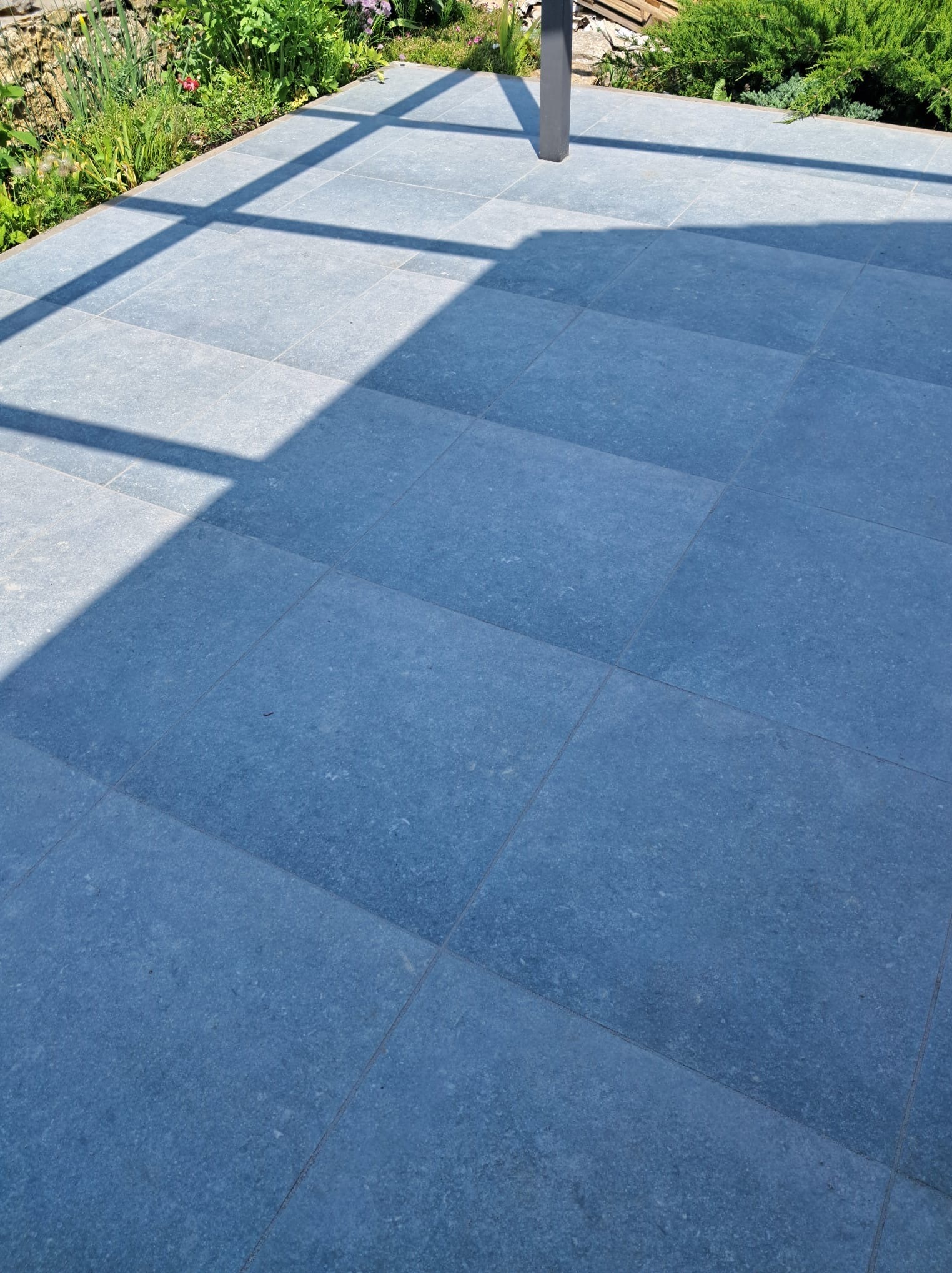 Niebiesko-szara, gładka posadzka z płytek na zewnątrz, oświetlona słońcem i cieniem konstrukcji. W tle zieleń roślin i fragmenty konstrukcji.