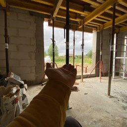 INSPIRATION BEAUTY Sp. z o.o. - Wnętrze w trakcie budowy z drewnianym szalunkiem stropu podpartym metalowymi stemplami. W tle widok na zewnątrz i dłoń osoby w kadrze.