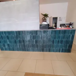 Bar z turkusowymi, pionowo ułożonymi płytkami i beżową podłogą. Na blacie ekspres do kawy i akcesoria. Ściana w odcieniach szarości.