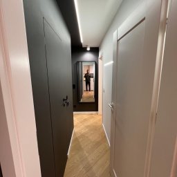 INSPIRATION BEAUTY Sp. z o.o. - Nowoczesny korytarz z parkietem w jodełkę, ciemną zabudową i lustrem, w którym odbija się postać. Oświetlenie LED podkreśla minimalistyczny design.