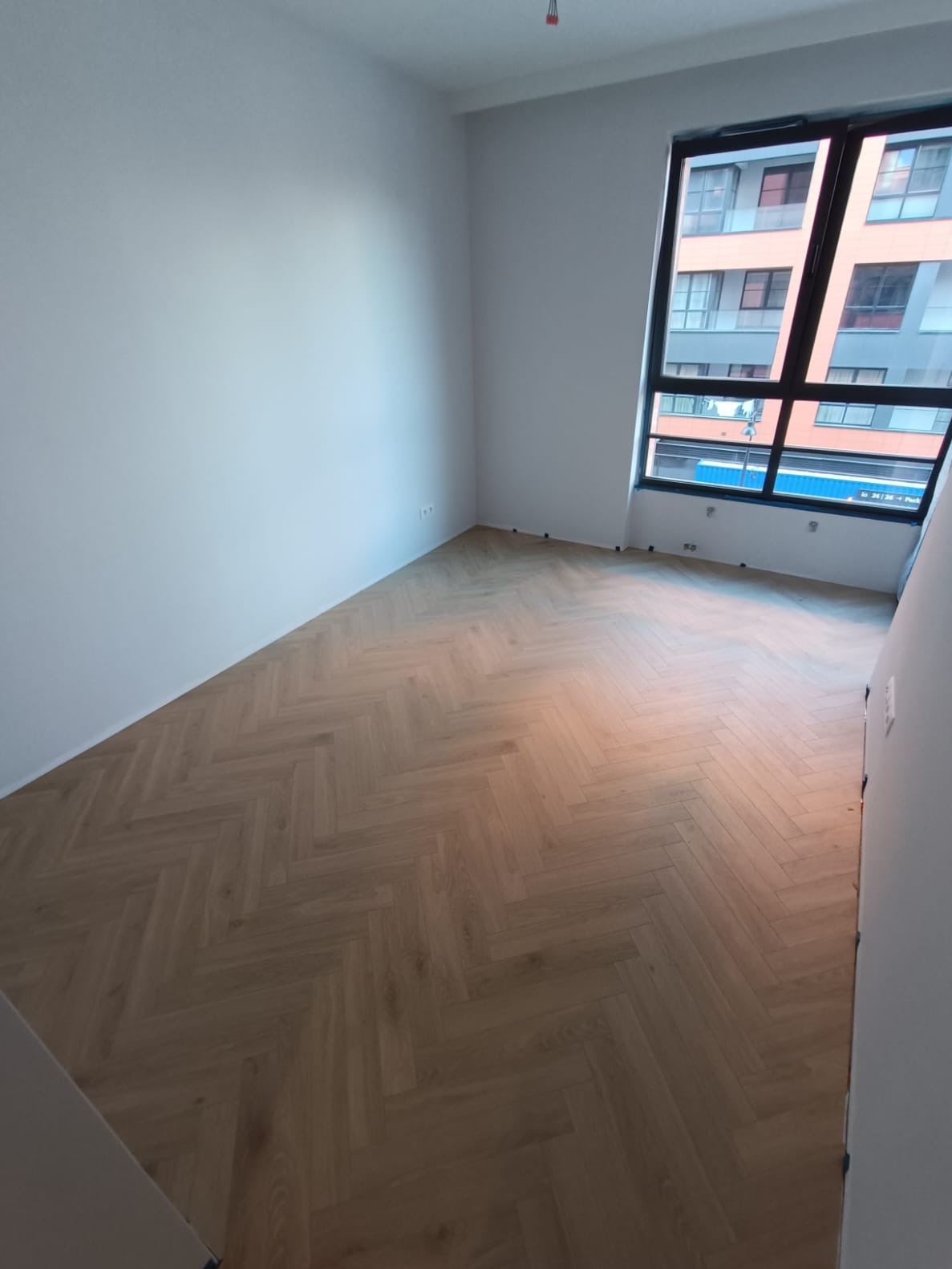 Jasne panele ułożone we wzór jodełki francuskiej w pustym pokoju z dużym oknem. Świeżo pomalowane ściany na biało. Widok na budynek z zewnątrz.