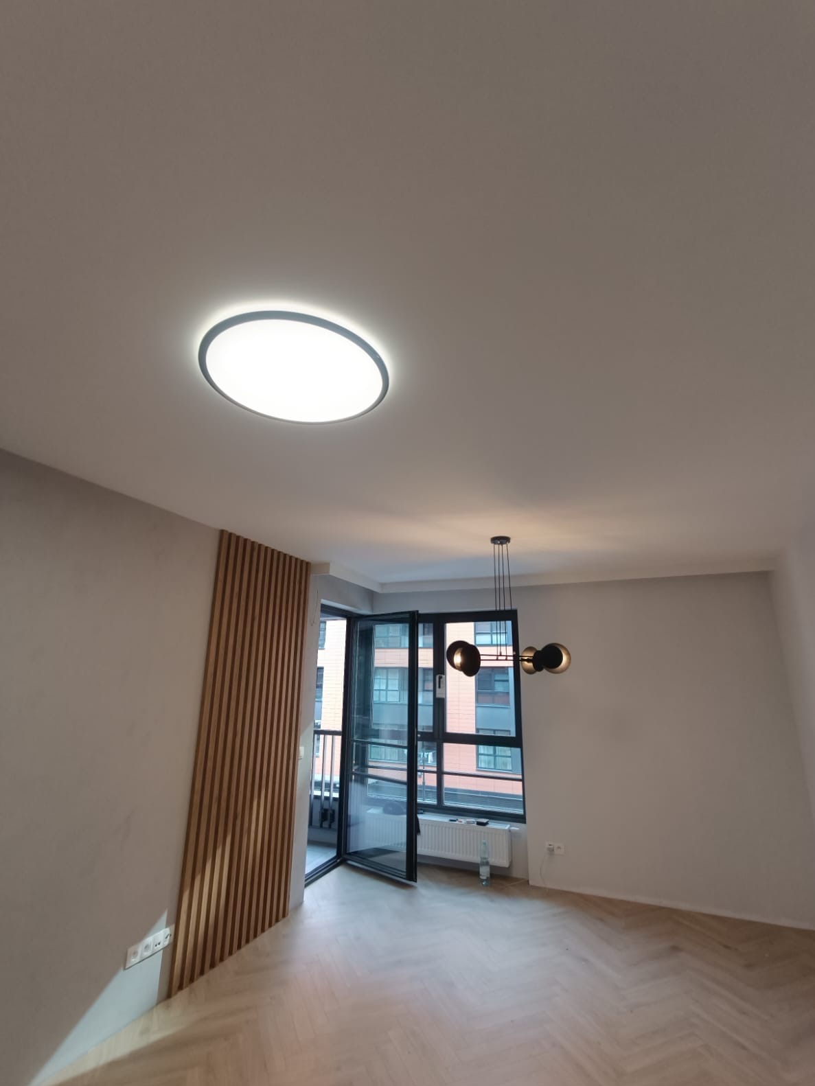 Jasne wnętrze po wykończeniu: parkiet w jodełkę, drewniana dekoracja ściany, nowoczesna lampa sufitowa i wisząca. Otwarty balkon z widokiem na miasto.