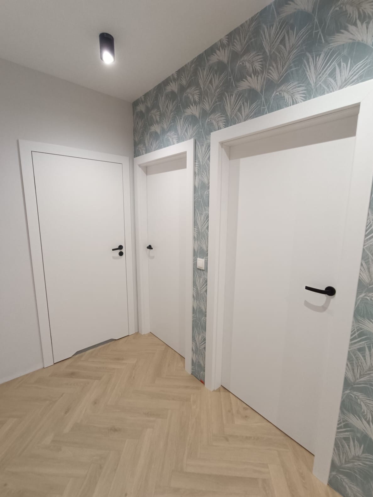 Korytarz z trzema białymi drzwiami z czarnymi klamkami, parkiet w jodełkę i ściana w tapetę z motywem liści. Nowoczesne wykończenie wnętrza.