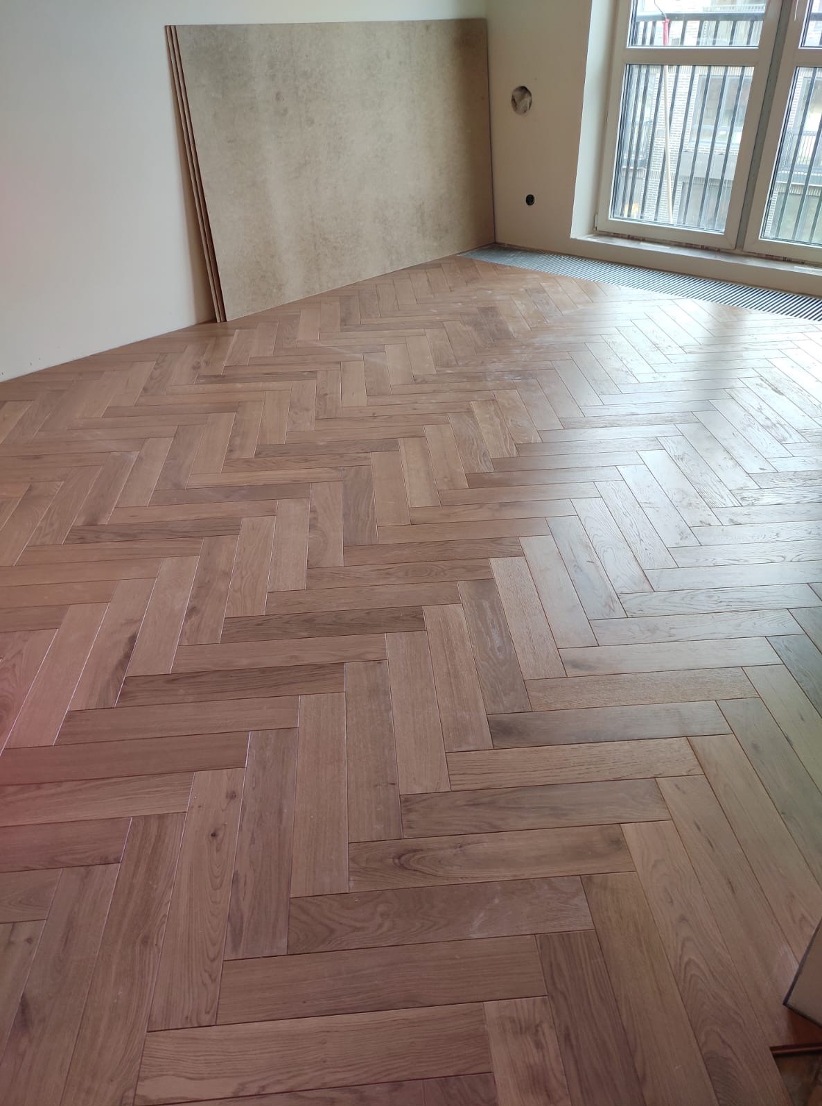 Ukończona podłoga z parkietu ułożonego w jodełkę, z widocznymi panelami przy ścianie i grzejnikiem pod oknem. Jasne, naturalne odcienie drewna.