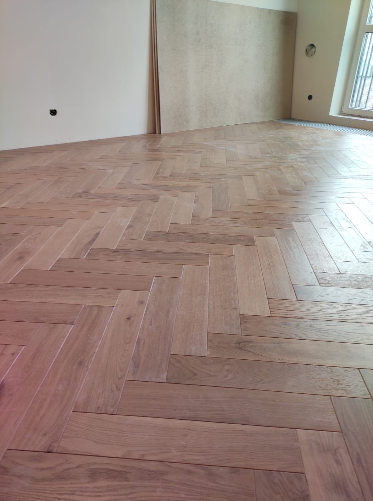 Parkiet ułożony we wzór jodełki w jasnym wnętrzu. Widoczne płyty kartonowo-gipsowe oparte o ścianę. Świeżo położona podłoga z drewna.