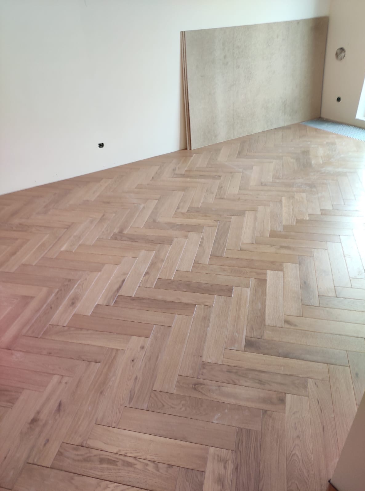 Ułożona parkietowa podłoga w jodełkę, jasne drewno, widoczne płyty gipsowe opierające się o ścianę, gniazdko elektryczne na ścianie, wnętrze w trakcie wykańczania.