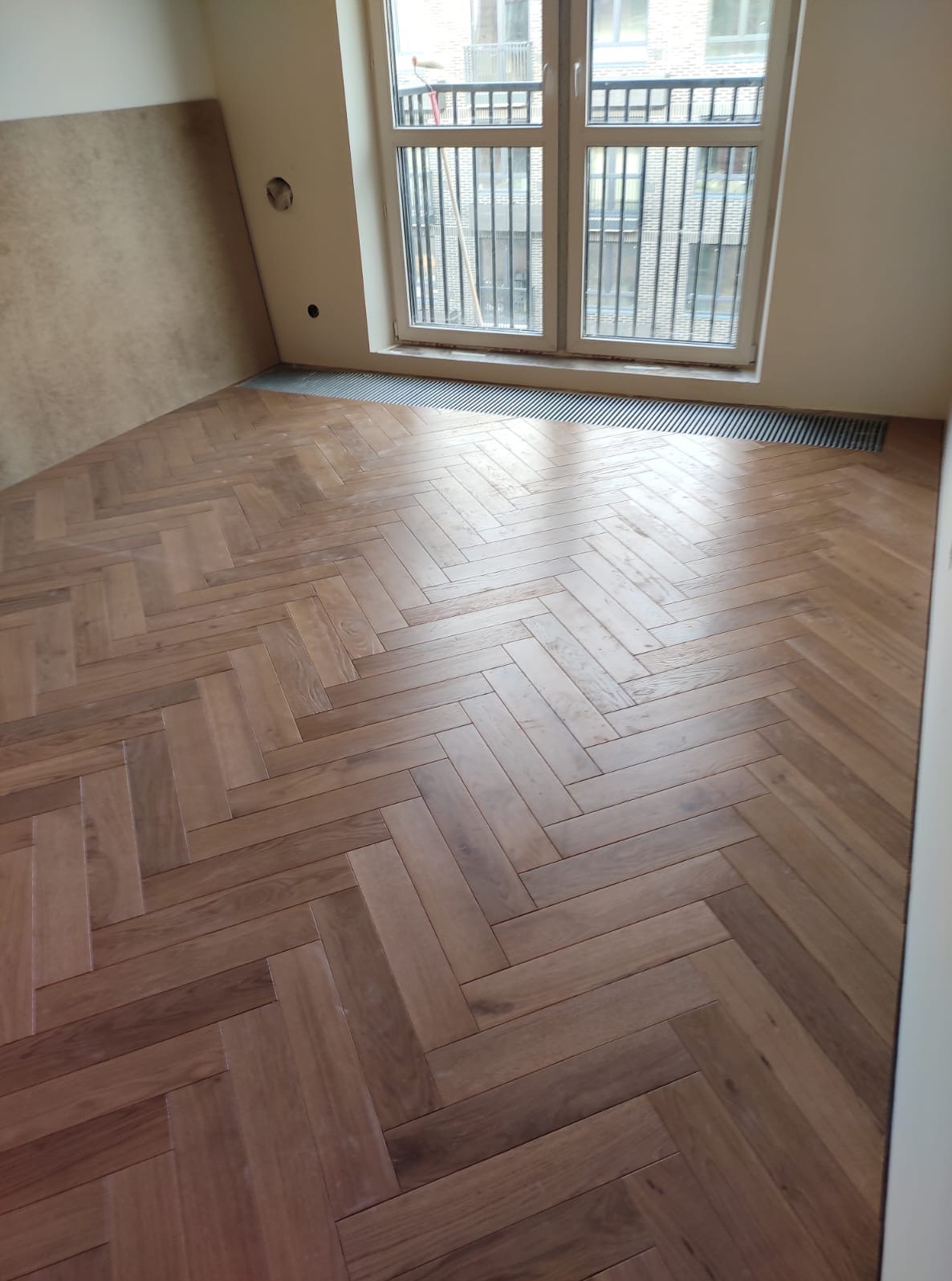 Parkiet ułożony we wzór jodełki w jasnym pokoju z oknem. Widoczne grzejniki pod oknem i fragment ściany przygotowanej pod dalsze prace wykończeniowe.