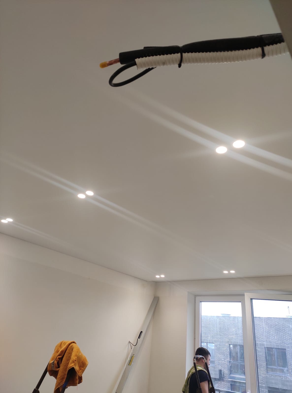 Biały sufit z zamontowanymi punktowymi lampami LED. Widoczna instalacja elektryczna w peszlu i fragment drabiny. W tle okno z widokiem na budynek.