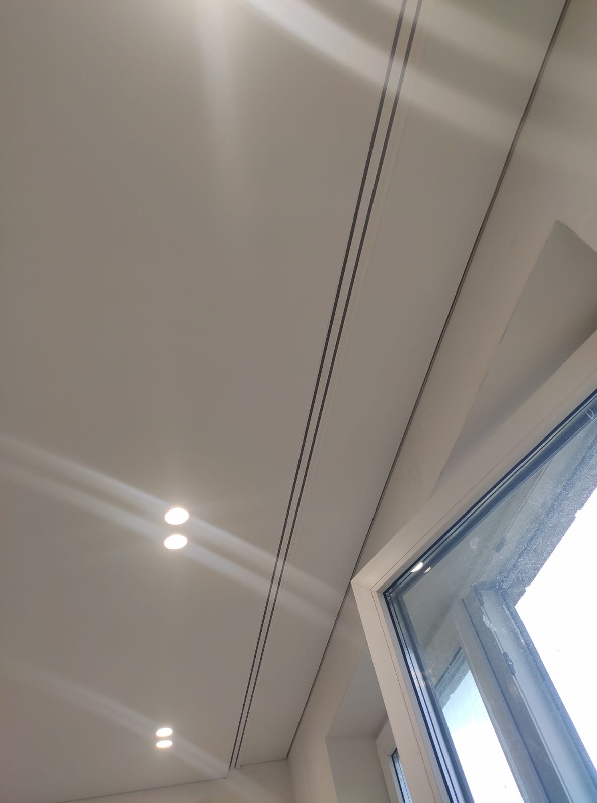 Biały sufit podwieszany z oświetleniem punktowym i czarnymi listwami dekoracyjnymi, widok z dołu, fragment okna po prawej stronie.