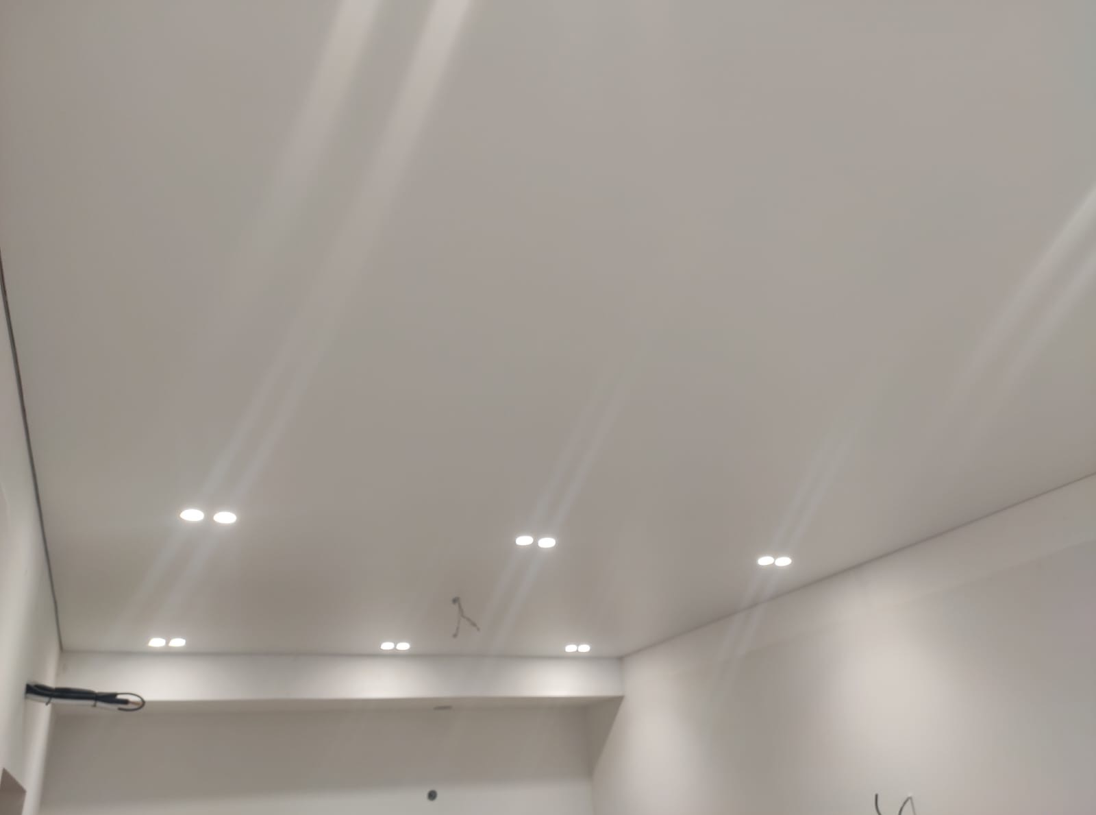 Biały sufit podwieszany z równomiernie rozmieszczonymi, punktowymi oprawami oświetleniowymi. Minimalistyczny design wnętrza, widoczne kable w narożniku.