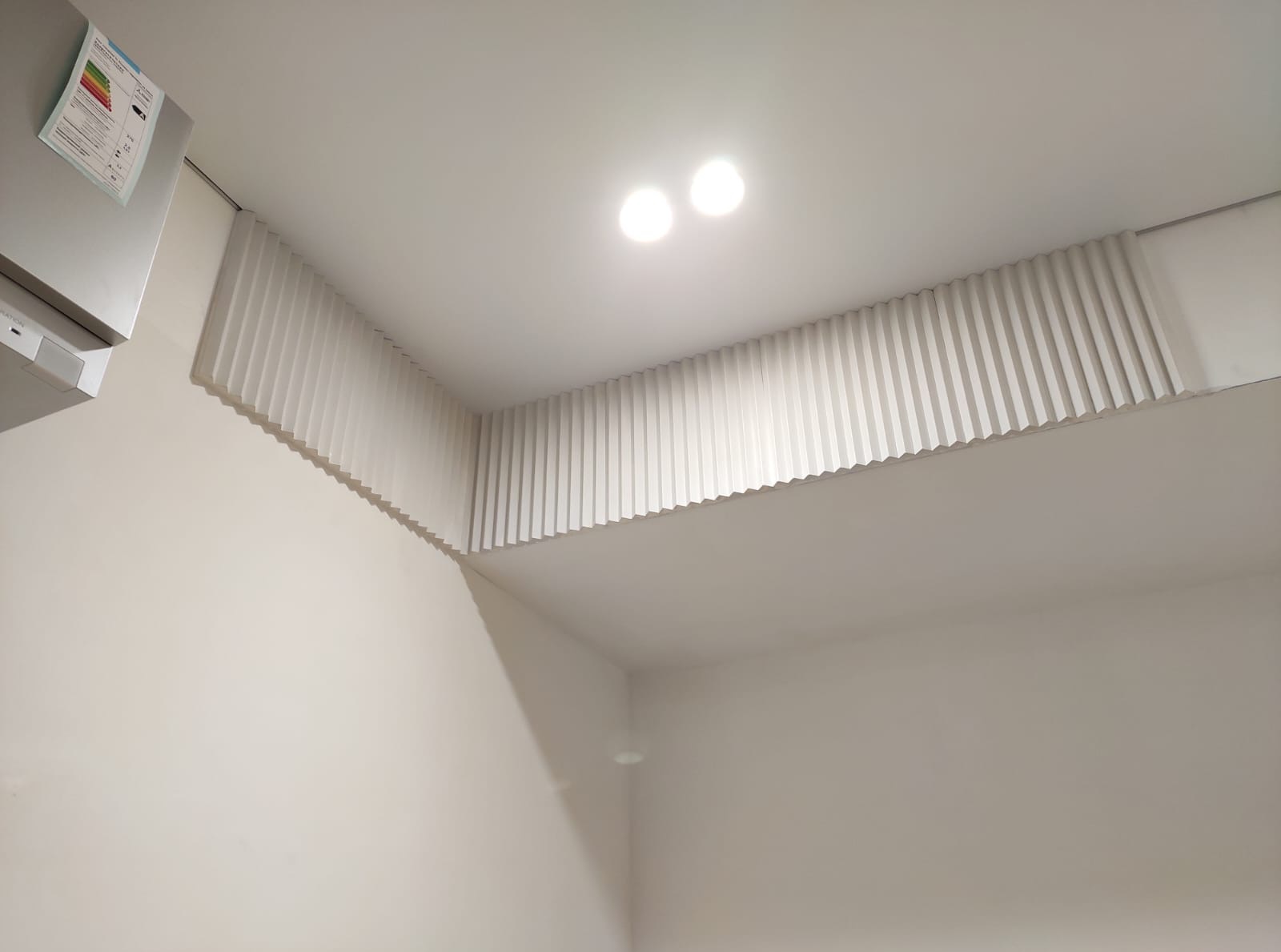 Nowoczesny sufit z dekoracyjnymi, pionowymi lamelami w jasnym wnętrzu. Widoczne oświetlenie punktowe i fragment urządzenia z etykietą energetyczną.