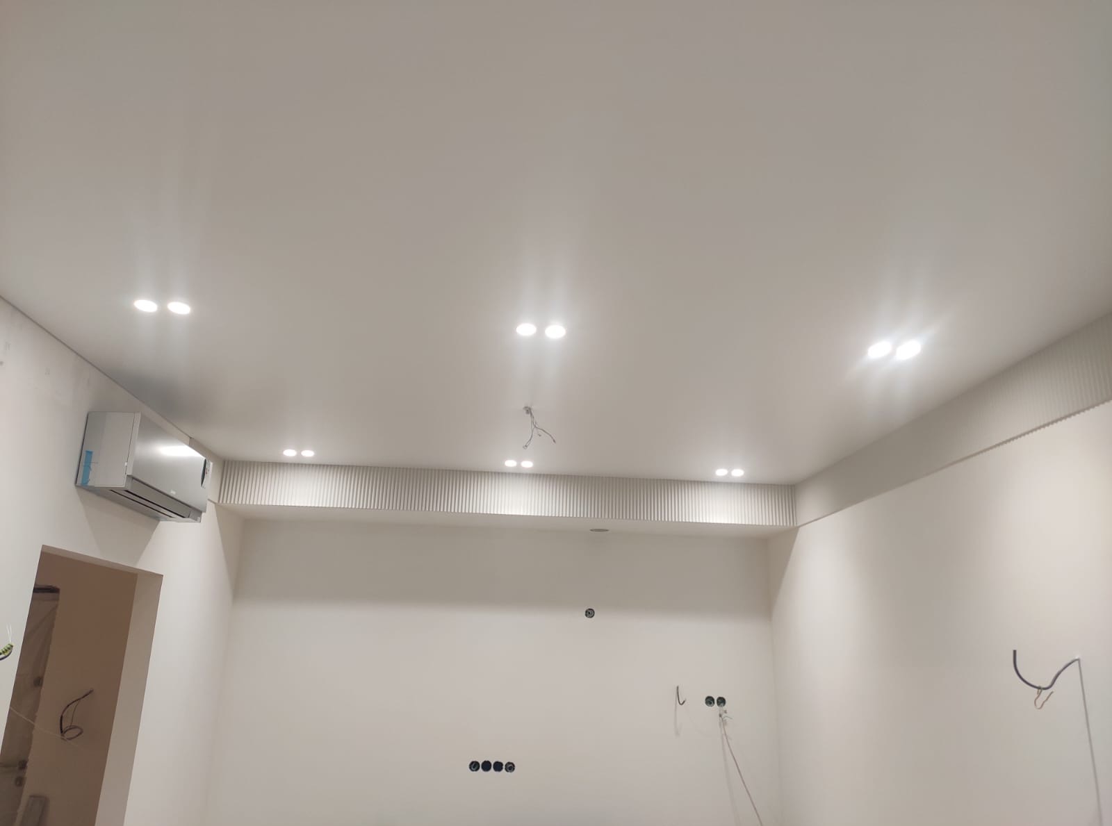 Białe wnętrze z klimatyzacją i oświetleniem punktowym w suficie podwieszanym. Dekoracyjne panele ścienne i widoczne kable sugerują prace wykończeniowe.