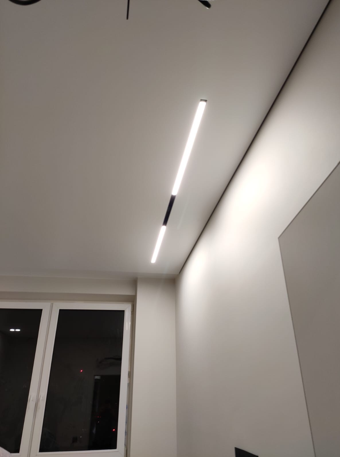 Nowoczesne oświetlenie sufitowe LED w minimalistycznym wnętrzu. Długa, smukła lampa z czarnym łącznikiem, równomiernie oświetlająca jasne pomieszczenie z oknem.