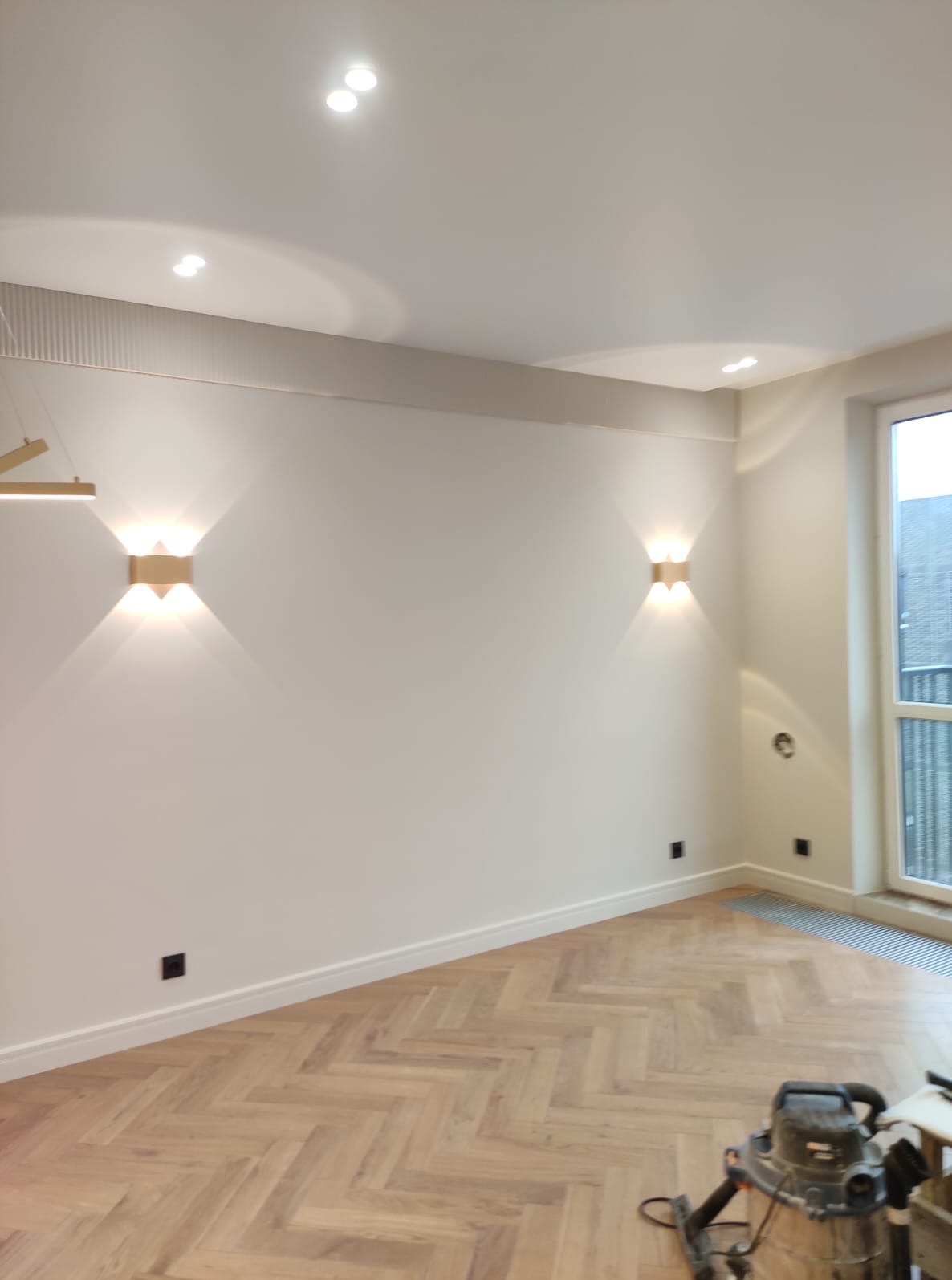 Jasne wnętrze po remoncie: parkiet w jodełkę, białe ściany z ozdobnymi listwami, nowoczesne kinkiety i punktowe oświetlenie sufitowe. Widoczny odkurzacz.