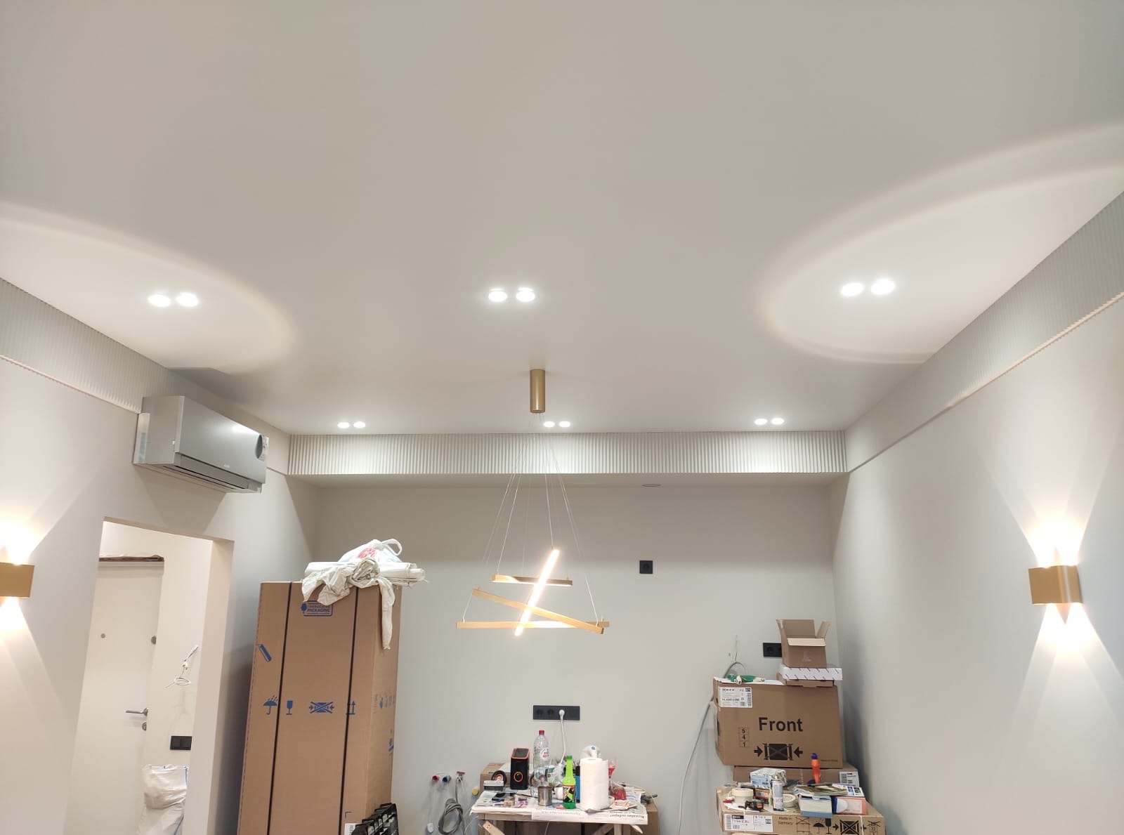 Nowoczesne oświetlenie sufitowe z klimatyzacją i dekoracyjnymi panelami ściennymi w jasnym wnętrzu. Widoczne kartony i akcesoria sugerują etap wykończenia.