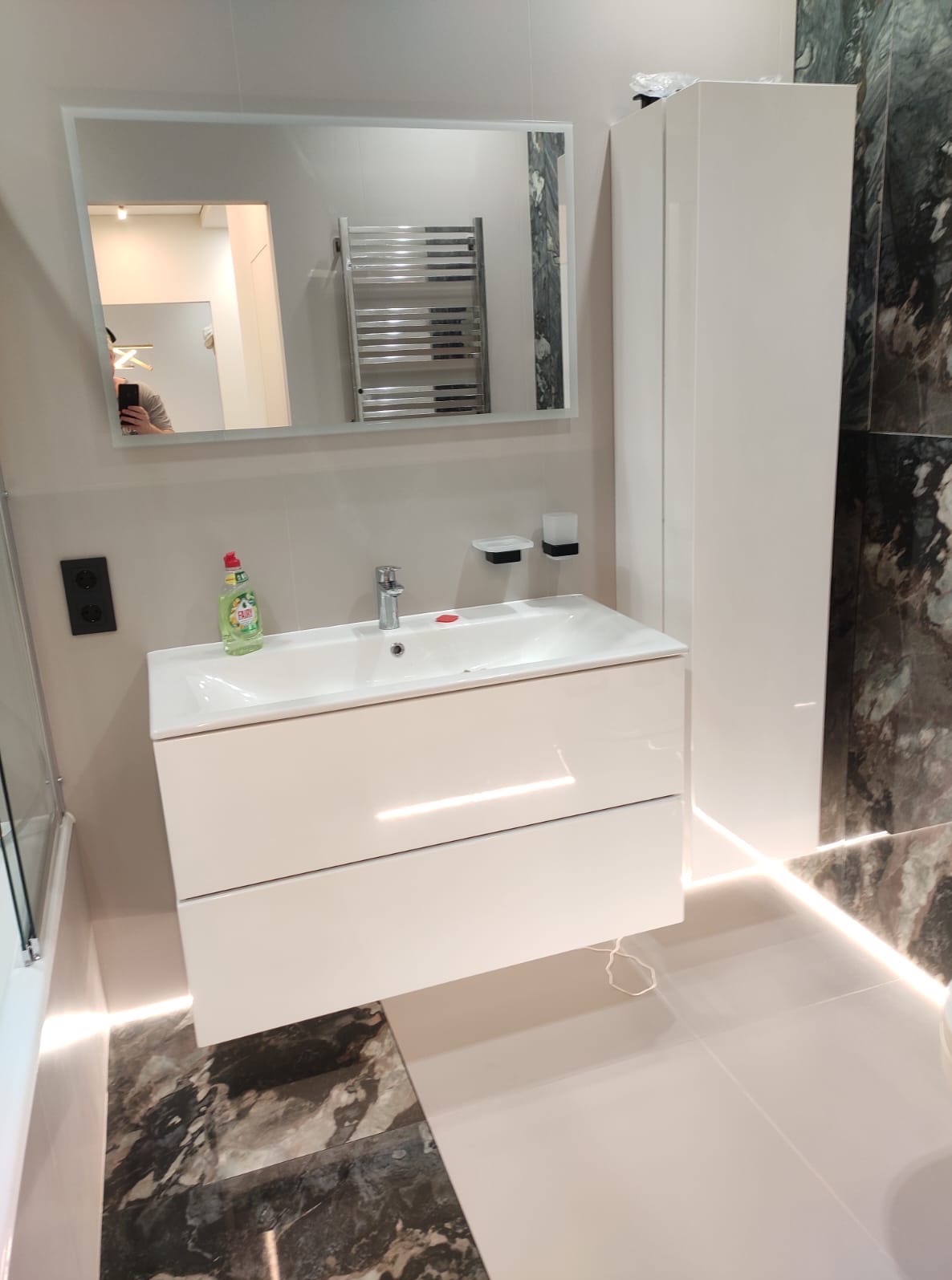 Nowoczesna łazienka z białą szafką pod umywalką, lustrem, grzejnikiem drabinkowym i marmurową płytką. Oświetlenie LED podkreśla elegancję wnętrza.