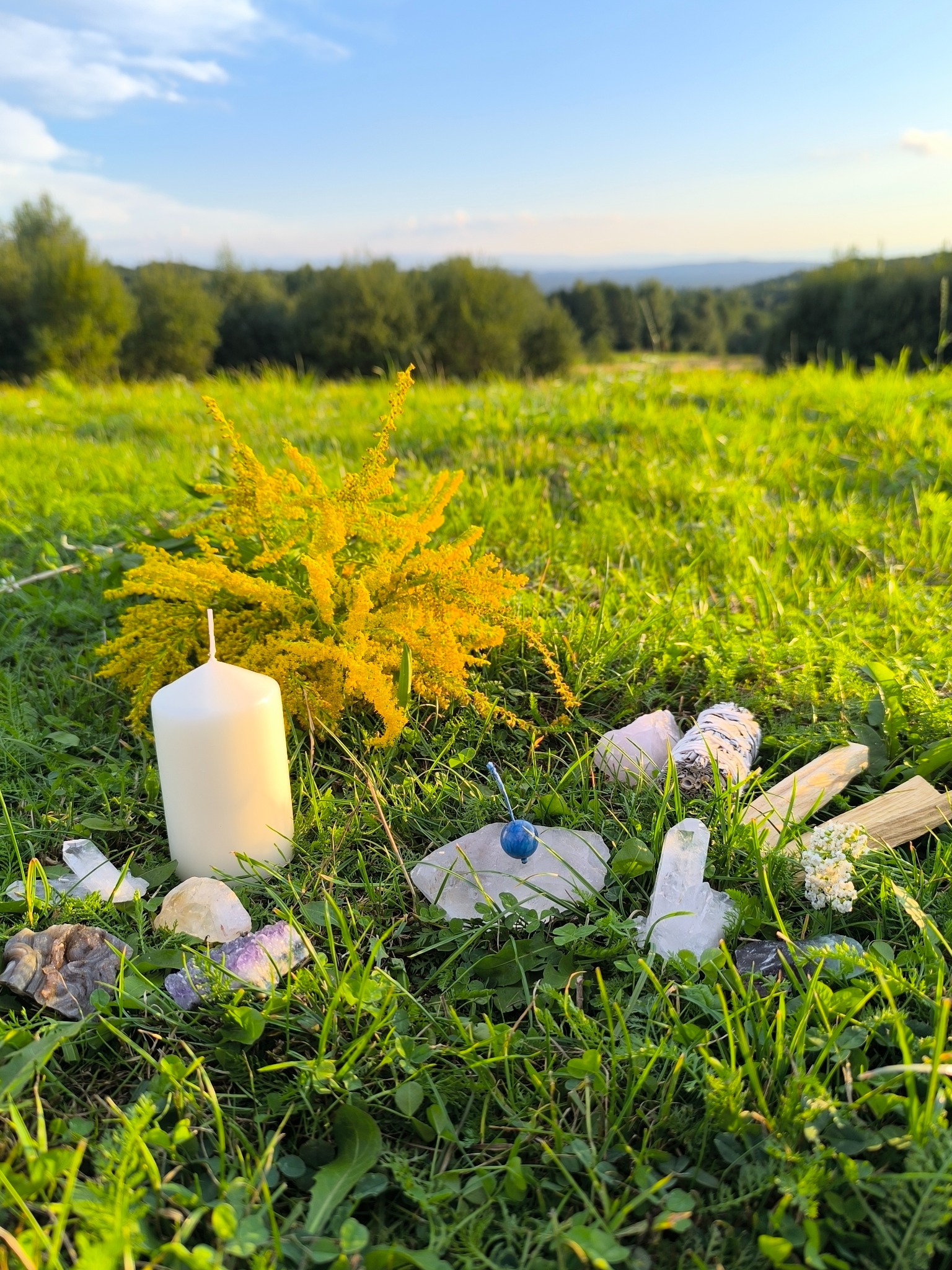 Świeca, szałwia, kamienie i żółty kwiat na trawie, w tle zielone wzgórza pod błękitnym niebem. Elementy do oczyszczania energii w naturalnym otoczeniu.