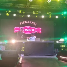 Zespół muzyczny na scenie z logiem 'Piekarnia AL Onkol'. Oświetlenie sceniczne w odcieniach zieleni, widoczni muzycy i instrumenty. Ujęcie z dołu, perspektywa widza.