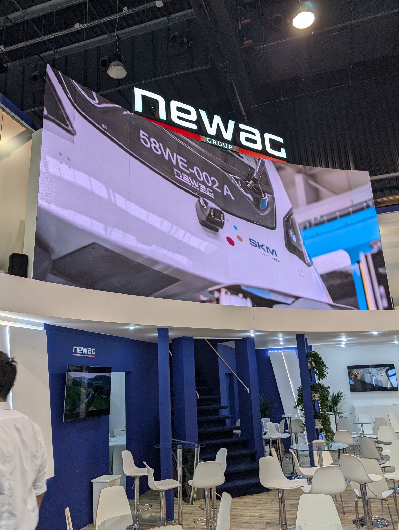 Stoisko targowe Newac Group z dużym ekranem LED prezentującym pojazd SKM. W tle widoczne stoliki i krzesła dla gości, schody i rośliny. Nowoczesny design.