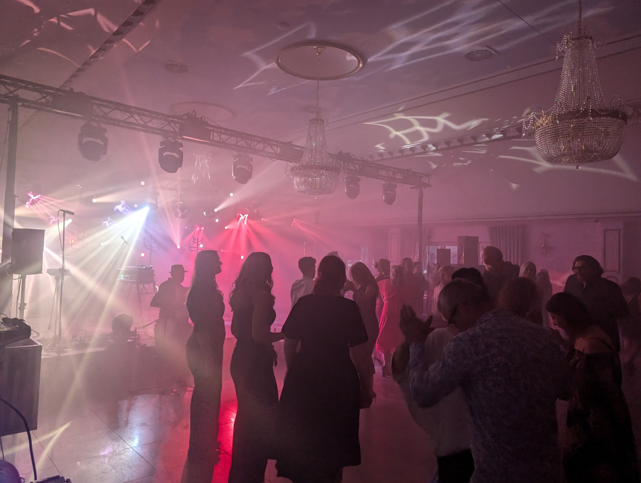 Sala balowa z parkietem, na którym tańczą ludzie w oświetleniu reflektorów i pod kryształowymi żyrandolami. Na scenie widoczny zespół muzyczny.