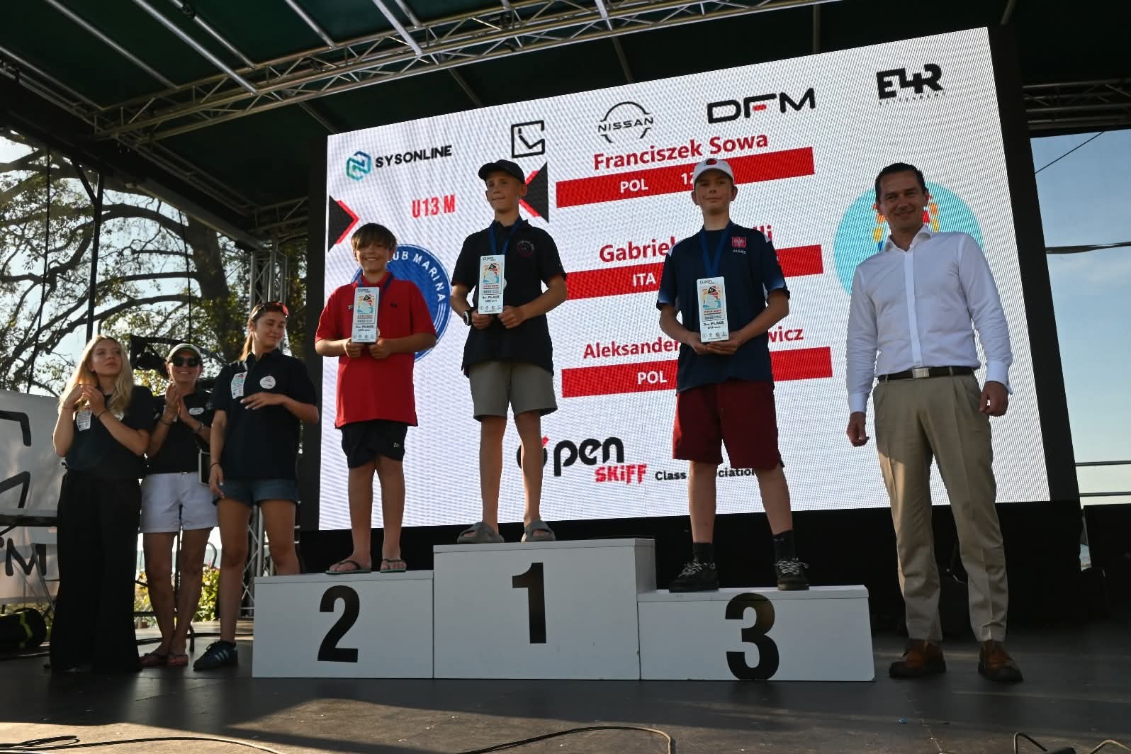 Uroczyste wręczenie nagród na podium dla młodych żeglarzy Open Skiff Class U13M. Sponsorzy na telebimie w tle. Osoby biją brawo. Widok z dołu.