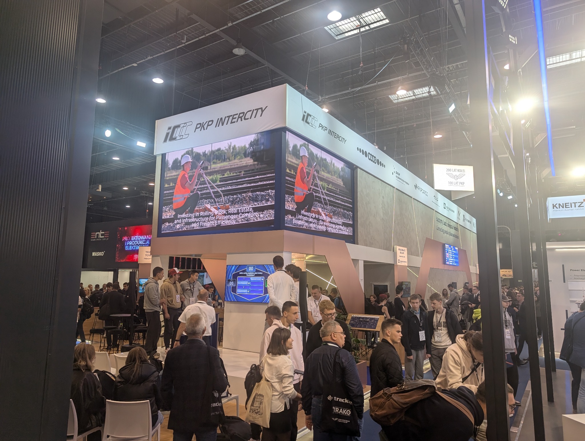 Tłum ludzi na targach, widoczny stoisko PKP Intercity z dużymi ekranami LED wyświetlającymi prace torowe. Oświetlenie jasne, ekspozycja targowa.