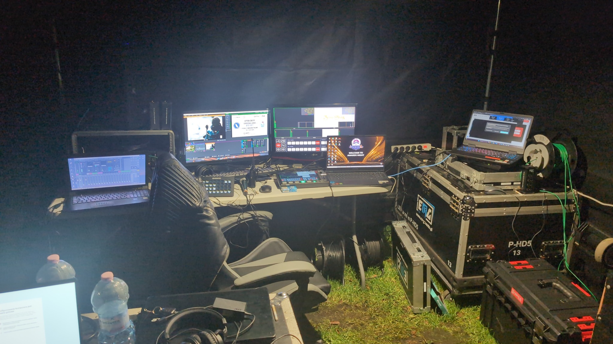 Stanowisko reżyserskie z kilkoma monitorami i laptopami podczas wydarzenia. Widoczne konsole sterujące, okablowanie i elementy nagłośnienia.