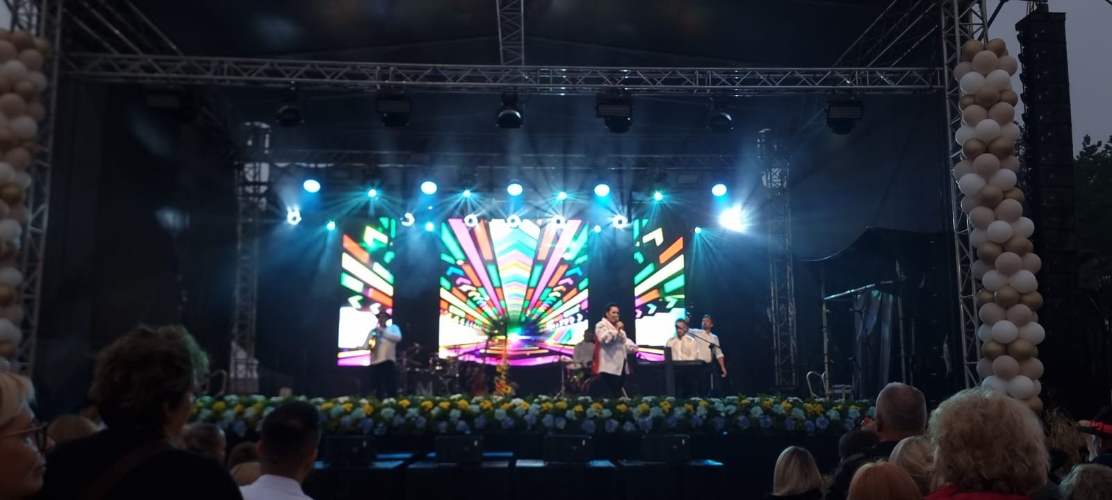Zespół na scenie z kolorowym ekranem LED, nagłośnieniem i dekoracją z kwiatów oraz balonów, widok z perspektywy publiczności.
