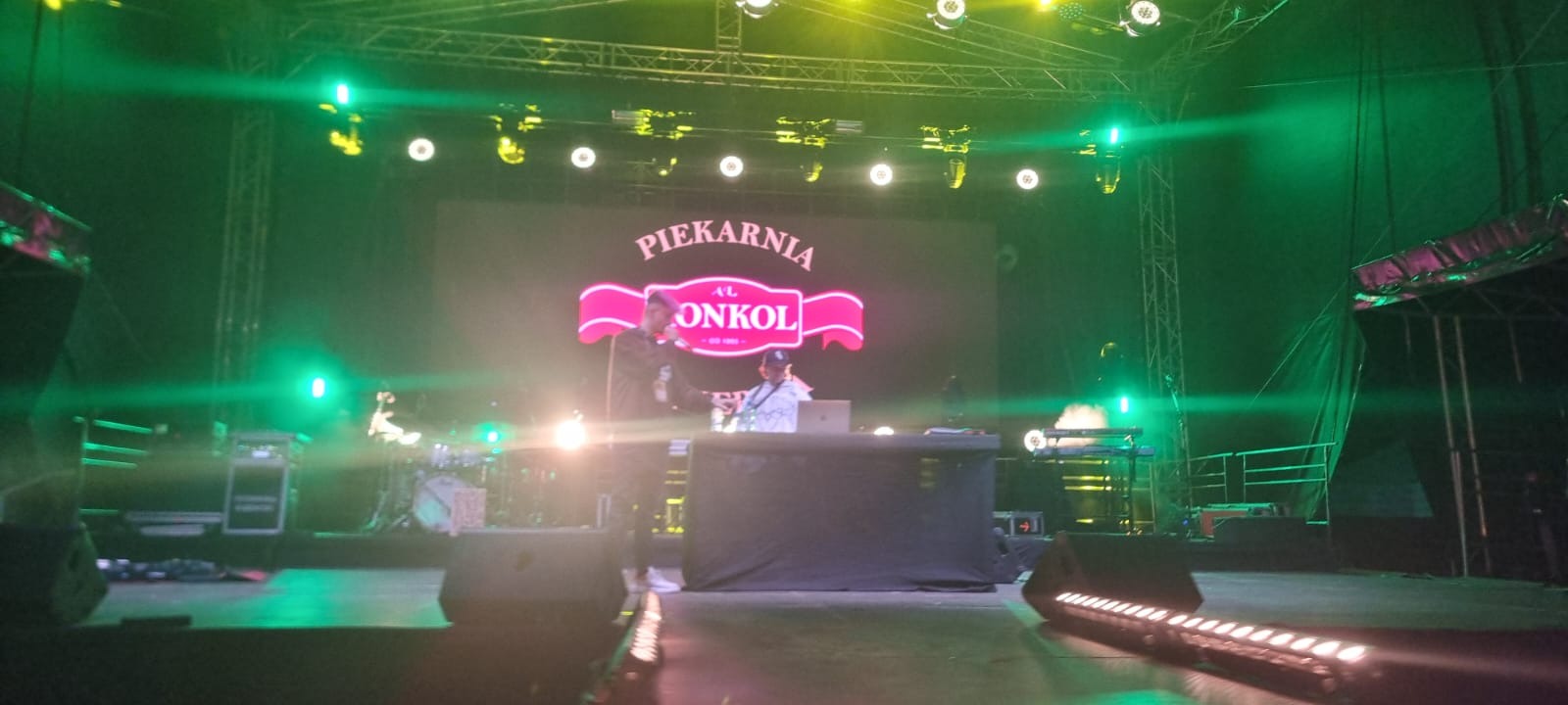 Zespół muzyczny na scenie z logiem 'Piekarnia AL Onkol'. Oświetlenie sceniczne w odcieniach zieleni, widoczni muzycy i instrumenty. Ujęcie z dołu, perspektywa widza.