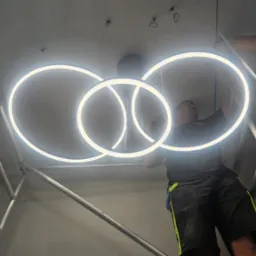 Elektryk na rusztowaniu montuje nowoczesną, okrągłą lampę sufitową LED z trzema pierścieniami. Widoczne kable i elementy mocujące.